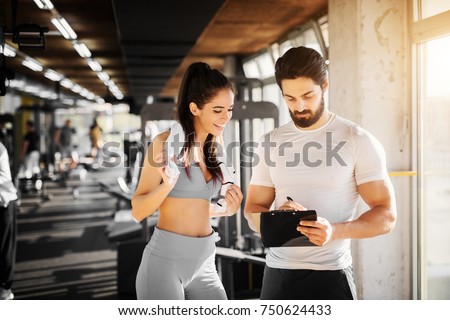 Young slim fitness girl standing with a towel near handsome trainer while showing her schedule for next week in the gym. Royalty-Free Stock Photo #750624433