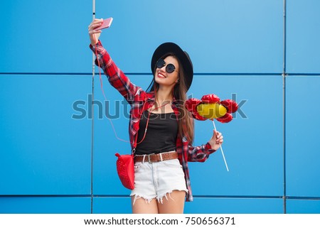 Fashion portrait of pretty smiling and woman in sunglasses with smartphone against the colorful blue wall. Make selfie with balloon in her hand