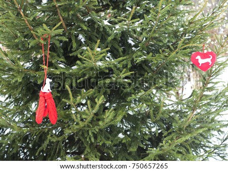 Paper dog figure on red decorative plush heart and red mittens hanging on fir tree green branches.