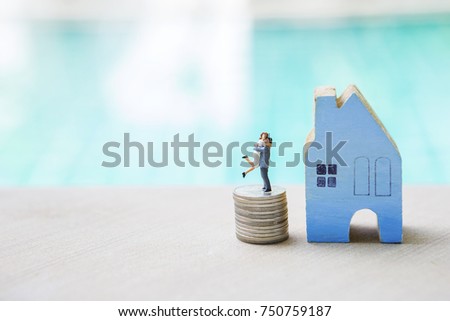 Miniature happy couple on stack of coin with wooden blue house over blurred blue swimming pool background, real estate and property business concept