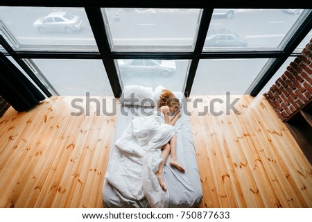 Young skinny beautiful barefoot girl lying in white bed with blanket and pillows and sleeping. Morning relax at sofa. Ginger woman resting in studio apartment with wooden floor and huge windows