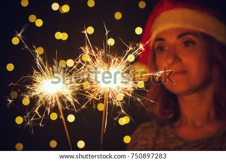 Holliday dynamic postcard. Young woman with a Santa Claus hat holds lighted fireworks in the glitter background with bright golden Christmas lights. Meeting the new year. Christmas party     