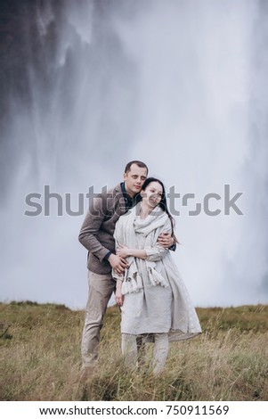 Iceland couple embrace near the big waterfall. Wall of the water on the background. Woman wear beautiful linen dress and knitted scarf. They are enjoy each other and nature around.