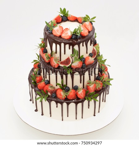 Three-tiered wedding cake in chocolate, decorated with slices strawberries, blueberries, figs and green leaves on a white background. Picture for a menu or a confectionery catalog.