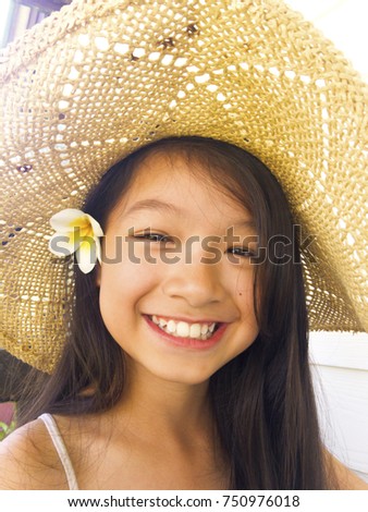 Asian long black hair girl is sitting on the wooden chair. She is smiling and wearing straw light brown hat and white flower behind the right ear