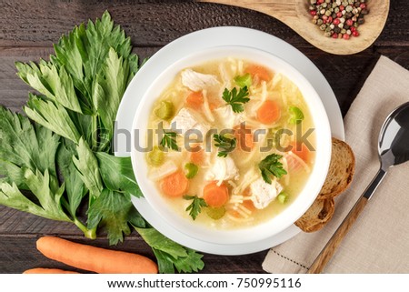 A closeup photo of a plate of chicken and noodles soup, shot from above on a dark rustic texture with a spoon, a wooden ladle with peppercorns, slices of bread, a celery branch, and a carrot