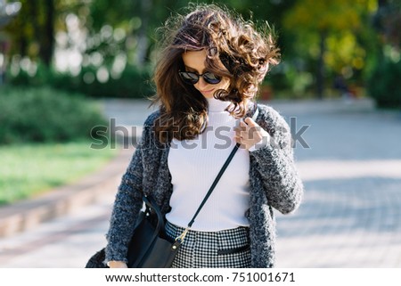 Trendy young woman with short curly beautiful hair is walking on the street in sunlight. She wears white shirt and grey pullover and black sunglasses. curly hair in tail is flying overhead from jump.