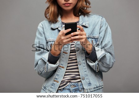 Cropped image of a serious young teenage girl dressed in denim jacket texting on mobile phone isolated over gray background