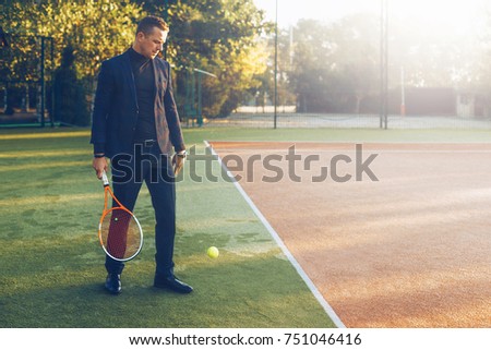 Portrait of confident businessman holding tennis racket in hand looking to jumping ball against tennis court background. A portrait of a tennis player wearing suit with a racket.