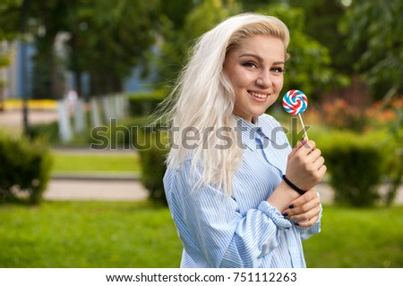 Outdoors portrait of beautiful blonde woman with candy in hands. Attractive student girl in summer sunny park.