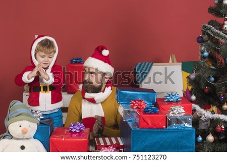 Man with beard and curious face plays with son. Christmas family opens presents on dark red background. Santa and little assistant among gift boxes near Christmas tree. Winter holiday concept