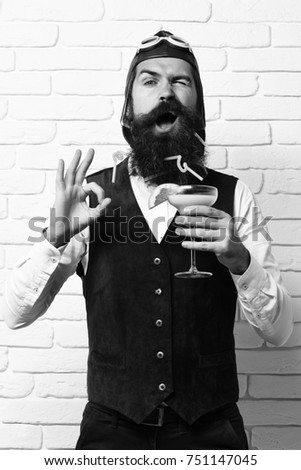 handsome bearded aviator man with long beard and mustache on funny face holding glass of alcoholic cocktail in vintage suede leather waistcoat with hat and glasses on white brick wall background