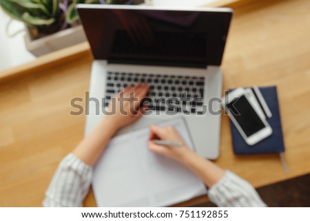 Top view of woman's hands typing on laptop keypad and writting in notebook placed on wooden desk with smartphone, credit card. Mock up. Blur image