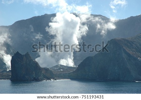 Smoke and steam is rising from the crater of an active volcano. The volcano is an island, and water is in the foreground. The sky is blue with no clouds.