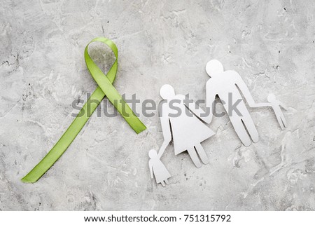 Green ribbon for Lyme disease, kidney cancer, organ donation awareness near paper silhouette of famoly on grey stone background top view