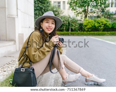 Beautiful Chinese woman sit at the edge of street, smiling with camera in hand, travel concept.