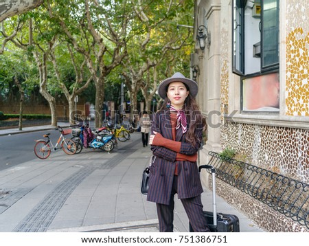 Beautiful Chinese woman in suit stand at the corner of street with suitcase beside her, arm crossed.