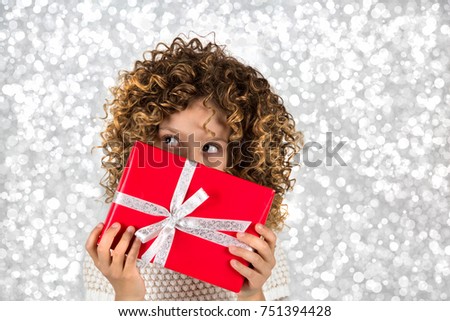 Red Gift. Picture of a little white caucasian girl with curly hair holding red gift box with white ribbon against bright white silver lights background