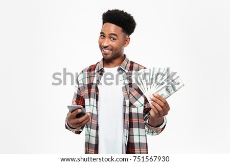 Portrait of a cheery young african man dressed in plaid shirt holding mobile phone and bunch of money banknotes isolated over white background