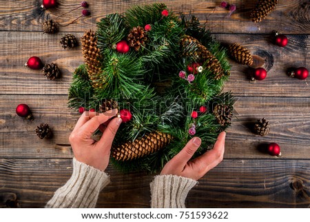 Preparation for xmas holidays. Woman decorating christmas green wreath with pine cones and red winter berries and christmas tree balls, wooden background, top view copy space, female hands in picture