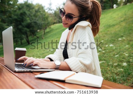 Picture of cheerful young woman wearing sunglasses sitting in park outdoors using laptop computer. Looking aside talking by phone.