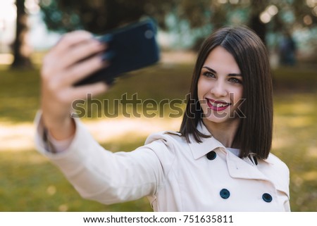 Close-up of beautiful young brunette taking selfie in park with smile on her face