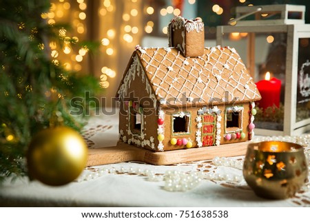 Little gingerbread house with glaze standing on table with tablecloth and decorations, candles and lanterns. Living room with lights and Christmas tree. Holiday mood Royalty-Free Stock Photo #751638538