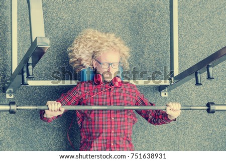Curly blonde guy in a red shirt trying to raise the barbell with effort. Toned