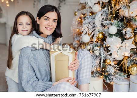 Thankful small female child embraces her mother who gave present, spend wonderful unforgettable time together, celebrate Christmas. Brunette woman and daughter look for present gifts under fir tree