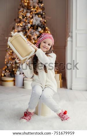 Vertical portrait of delightful small female child in white clothes and warm socks holds big present box, receives gift from parents on New Year, poses against decorated Christmas tree