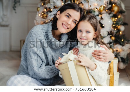 Cheerful brunette female leans at her daughter, embraces her, presents gift box, being in living room near decorated New Year tree. Glad family mother and daughter in warm sweaters celebrate Christmas