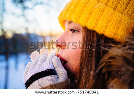 Hipster girl in yellow cup freezing in winter park in snowy sun winter day. Woman blowing on freezed hands wth coloful mittens. Pictures in warm colors. Close up, side view