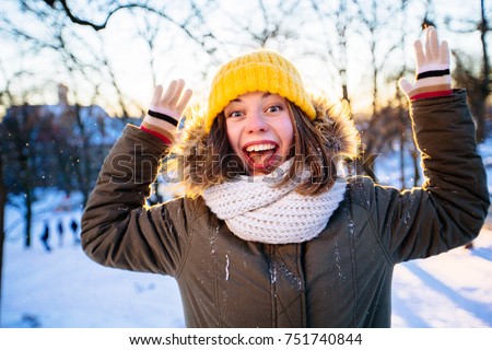Hipster crazy girl with open mouth in yellow cup heaving fun in winter park in snowy sunny winter day. Woman balancing and wave with her arms wth coloful mittens. Pictures in warm colors.