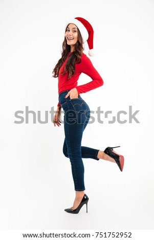 Side view full length portrait of a smiling cheerful asian woman dressed in red santa hat posing while standing and looking at camera isolated over white background