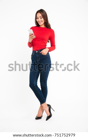 Full length portrait of a happy satisfied asian woman texting on mobile phone while standing isolated over white background