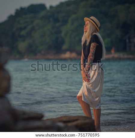 Beautiful young blond woman in straw hat walking on the morning beach on the background of turquoise water. Modern girl in a long skirt.