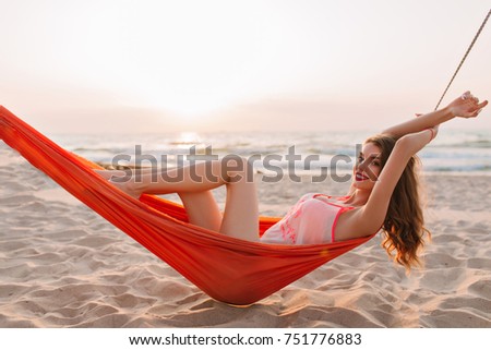 Stunning european female model in swimsuit posing with pleasure in hammock. Outdoor photo of happy young woman relaxing at ocean resort with sunset on background.