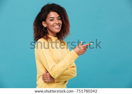 Photo of happy young african lady dressed in warm sweater standing isolated over blue background. Looking camera while pointing.