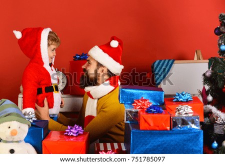 Man with beard and smiling face plays with son on New Year eve. Boxing day and family concept. Christmas family opens presents on red background. Santa and little assistant among gift boxes