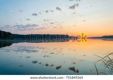 A calm dawn on a mirror lake with a forest overlooking the sun on the horizon