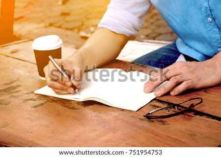 In the morning . Men sitting in the park  and he writing notebook. He drinking coffee and working on idea on notebook in relax time .
