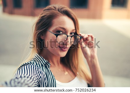 Half length portrait of gorgeous positive blogger looking at camera and correcting spectacles while walking at urban setting.Charming hipster girl in good mood spending leisure time at street