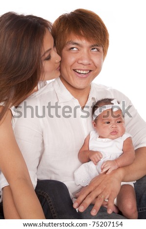 A mother kissing her husband on the cheek while he is holding their baby girl.  The father has a funny expression on his face.