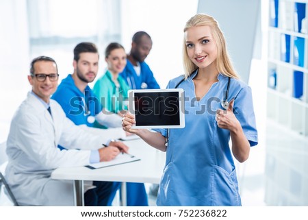 young nurse holding tablet and showing thumb up with colleagues sitting at table on background