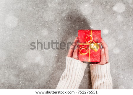 Preparing for the holidays, Christmas background. Female hands in the picture in a warm sweater hold a gift in a red wrapper with a golden ribbon. Gray background, snow effect, Top view copy space