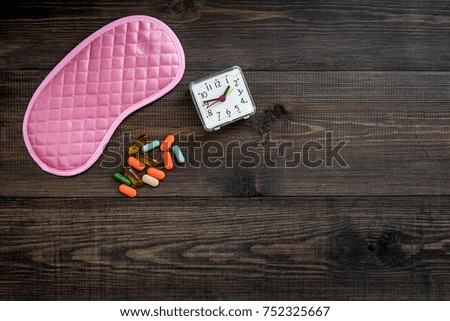 Sleeping pills for insomnia near sleep mask and alarm clock on dark wooden background top view.