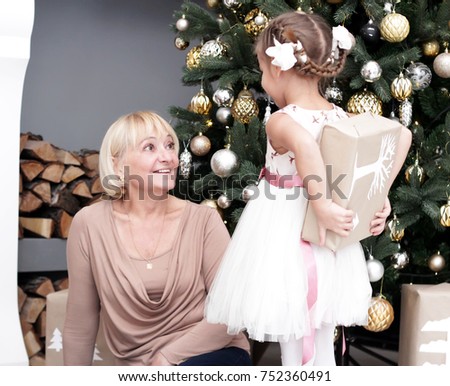 Portrait of happy granddaughter hiding Christmas present for her grandmother behind her back, happy holiday family over decorated New Year tree, merry Christmas concept, lifestyle indoor, studio shot