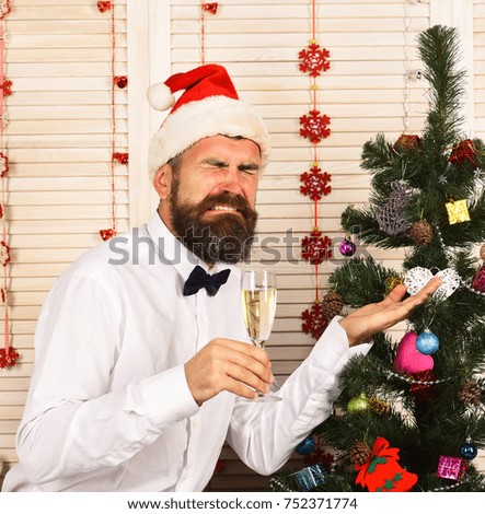 Celebration and New Year concept. Guy near Christmas tree on wooden wall background. Santa Claus in red hat with grimace. Man with beard and bow tie holds champagne glass and shows fir tree