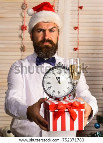 Man with beard holds striped present box with champagne glass and alarm clock. Santa Claus in hat with strict face. Guy on wooden wall and garlands background. Celebration and surprise concept