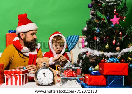 Santa and little assistant among gift boxes near Christmas tree. Family holidays concept. Man with beard and curious face play with son. Christmas family opens presents on green background